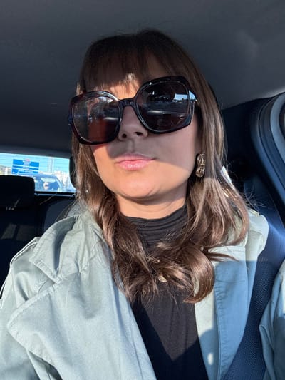 A brunette woman wearing large sunglasses and gold earrings poses. She has on a black turtleneck and a light gray jacket. She is seated in what appears to be a car. Light streams in, casting shadows on her face and clothing. Part of the car's interior is visible in the background.