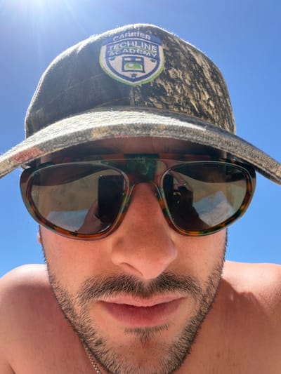 A man wearing a bucket hat and sunglasses looks directly at the camera. The hat has a "Carrier Techline Academy" patch. His dark facial hair contrasts with his fair skin. The sky appears bright blue in the background. The sunglasses reflect the surroundings. The photo is angled slightly downwards.