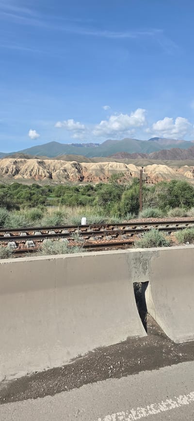 The image depicts a landscape scene in Krasnyy Most, Kyrgyzstan. In the foreground, a concrete barrier with a triangular opening runs horizontally across the frame, separating the viewer from the rest of the scene. Beyond the barrier, a set of train tracks is visible, with rusting rails and concrete sleepers partially obscured by green vegetation. Further in the background, a dense line of green trees and bushes stretches across the mid-ground. Behind the vegetation, a series of layered hills with stratified rock formations in shades of tan and reddish-brown dominate the landscape. The distant background is characterized by rolling green mountains, with some peaks showing patches of snow under a bright blue sky with scattered white clouds. The overall impression is one of a rural or semi-rural environment with evidence of infrastructure.