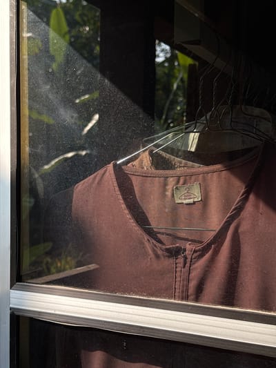 A close-up shot shows a brown shirt hanging on a wire hanger. The shirt has a zippered neckline. A label with a hanger icon and some text is visible on the shirt. The shirt is viewed through a glass window, and the window is dusty, with streaks of light from the sun shining through it. Outside the window, greenery and foliage are visible. The scene is likely located in Kalirancang, Indonesia, given the context of the city.