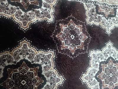The image displays a close-up, high-angle view of a dark brown patterned textile, located in Dashtabad, Uzbekistan. The fabric exhibits a repeating design featuring intricate motifs in off-white, light brown, and dark brown. Prominent elements include stylized multi-pointed star or rosette medallions, primarily in off-white with detailed brown internal patterns, set against a textured, deep brown background. These medallions are bordered by arc-shaped segments containing smaller scrollwork and dotted lines. The textile surface appears plush, consistent with a carpet or velour material, showing slight variations in nap. No individuals, other objects, or actions are visible within the frame.
