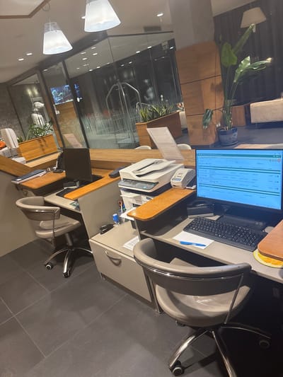 The image captures an indoor office or reception area, likely within a commercial establishment in Dobrinishte, Bulgaria. The space is unoccupied, presenting an orderly and functional atmosphere typical of a front office or administrative workstation.

The central feature is a desk area comprising a wooden surface and grey filing cabinets. A computer monitor displays what appears to be a booking or inventory management system, showing a tabular interface with columns labeled "Item," "Event," "Date," "Status," and "Price." Visible entries include "Test Reservation," dates like "13.08.2021," "02.04.2023," and "28.02.2023," alongside prices, including "115.00 EUR." A search bar reads "Search reservations / guests..." A keyboard, mouse, and a blue pen on a mousepad are positioned in front of the monitor. Adjacent to the desk, a white multi-function printer/scanner is visible, and on a lower shelf, various office supplies such as remote controls, scissors, and a pen holder are organized. A grey upholstered desk chair with a chrome frame is placed at the workstation, and a distinct orange-colored circular coaster or decorative item rests on the desk.

Adding a natural element to the interior are large potted plants with broad green leaves, possibly a Bird of Paradise, situated behind the main desk. The background features wooden wall panels or cabinetry, and a reflective glass partition offers a glimpse into another interior space. Two modern white conical pendant lights illuminate the area from above. The floor is tiled with large, dark grey squares. A partial view of another similar workstation with a monitor and chair can be seen in the lower foreground. The overall impression is one of a well-maintained and ready-to-use professional environment.