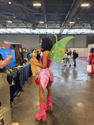 In the city of Villepinte, France, inside a large indoor convention center, a Black woman is seen from the chest down wearing a pink, strapless fairy costume with large green fairy wings. She has on knee-high pink platform boots. Her arms are bent at the elbows, and she is holding a red heart-shaped item in her right hand. In front of her, to the left, a person wearing khaki pants and a dark blue shirt is partially visible from the waist down, holding out their left arm, which has a pink and green floral tattoo. To the left of the woman in the fairy costume, a display table is set up with a blue tablecloth. On the table, there is a monitor displaying a blue architectural scene, several bouquets of flowers, and a tall grandfather clock. In the background, other convention attendees can be seen, including one person dressed as Ladybug from the show Miraculous. The floor of the convention center is made of concrete. The ceiling has exposed metal beams and fluorescent lighting.