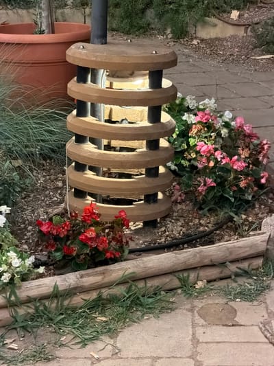 In the city of Cavriglia, Italy, a unique, cylindrical garden light fixture stands illuminated in the center of the frame. The fixture is constructed from stacked, circular wooden elements, each grooved with concentric rings, and supported by vertical black poles. Within the fixture, a light bulb emits a warm glow. The light is surrounded by a bed of mulch, stones, and various plants. To the left, a large, terracotta planter holds a small tree. In front of the planter, a cluster of thin, green grass grows. To the right of the light fixture, several clusters of flowering plants, featuring bright pink and white blossoms, add vibrant color to the scene. In the foreground, a raised garden bed is formed by two horizontal, weathered wooden logs, with green grass growing between them and along the edge of a paved walkway. The ground is paved with rectangular, stone tiles. A black hose or cable snakes across the ground behind the wooden logs. The scene appears to be an outdoor garden or patio area.