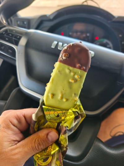 A close-up, eye-level shot captures a person's hand holding a Magnum ice cream bar inside the cabin of a vehicle. The ice cream bar is distinctive, featuring a vibrant green base, likely pistachio flavored, generously topped with a dark brown chocolate coating embedded with small nut pieces. The golden foil wrapper, partially peeled down, is visible at the bottom of the ice cream, displaying the brand name "MAGNUM" and the text "NEW CRACKING SHELLS". Other partial words like "GREEN" and "SHELLS" are also discernible on the wrapper.
The hand holding the ice cream has visible skin texture and short fingernails, with a faint dark marking or tattoo on the wrist. In the background, the interior of what appears to be a truck or commercial vehicle is discernible. The steering wheel, prominently featuring the "IVECO" logo, occupies much of the upper left and center-right of the frame. Beyond the steering wheel, the vehicle's dashboard is softly blurred, but some red and a green indicator light from the instrument panel are visible. The bright lighting suggests it is daytime. A blurry glimpse of a leg or foot is visible in the lower right corner, indicating the presence of a passenger or driver. The scene is set within a vehicle in Torrent, Spain.