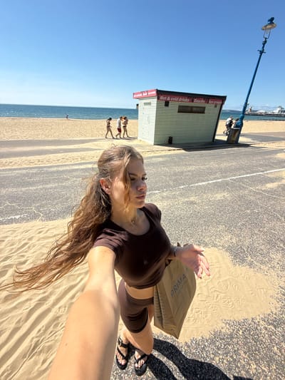 The image depicts a sunny day on a sandy beach in Bournemouth, United Kingdom. A young woman with long, wavy brown hair is in the foreground, holding a Primark shopping bag and extending her right arm, as if taking a selfie. She is wearing a brown crop top and matching shorts. Behind her, the beach stretches out to the sea under a clear blue sky. Several people are visible walking on the sand near a small, light-green beach hut with red signage indicating "Hot & cold drinks," "Snacks," and "Ice cream." A tall, blue lamp post stands to the right of the hut, leaning precariously. The ground in the foreground is a mix of sand and asphalt.
