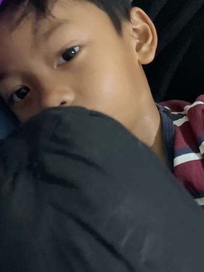 The image is a close-up, slightly tilted shot of a young child lying down. The child's face is partially obscured by dark fabric, but their eyes, nose, and ear are visible. The child appears to be wearing a striped shirt with maroon, white, and dark blue stripes. The background is dark and appears to be a soft surface, possibly a bed or sofa. The lighting suggests an indoor setting, possibly in the evening or with artificial light. The child's expression is neutral, and the overall mood of the image is intimate and casual. There is no visible text in the image. The location context provided is Serdang Kulon, Indonesia.