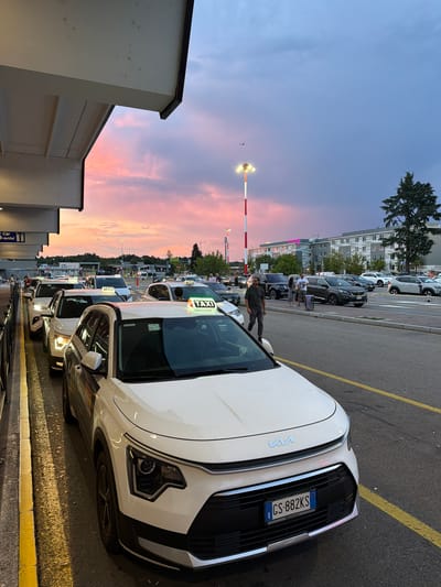 A queue of white taxis, many with illuminated "TAXI" signs, are parked on a roadside in front of a building. The sky is a gradient of twilight colors, transitioning from pink and orange near the horizon to deep blue. A tall light pole with red and white stripes stands to the left of the frame, and an airplane is visible in the distance. People are walking along the sidewalk next to the taxis, and parked cars are visible in the background. The scene appears to be at an airport or transportation hub in Somma Lombardo, Italy, given the presence of taxis and the airport lighting.