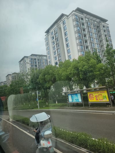 The media file depicts an outdoor scene in Yufengshan, China. The primary subjects are two multi-story buildings with numerous windows, suggesting a residential or office complex. Lush green trees are interspersed between the buildings and along the roadside, indicating a well-maintained urban or suburban environment. A paved road with white lane markings runs parallel to the buildings, and a sidewalk is visible. On the sidewalk, there are two billboards. The billboard on the left is yellow with red Chinese text and images of food products. The billboard on the right is blue and white with what appears to be informational graphics or text. To the right of the road, there is a parked scooter with a reflective shield attached to its handlebars. A person is partially visible seated on the scooter. The weather appears overcast, contributing to a muted color palette. The overall mood is calm and ordinary, suggesting a typical day. The scene is captured from a slightly elevated perspective, possibly from a vehicle.