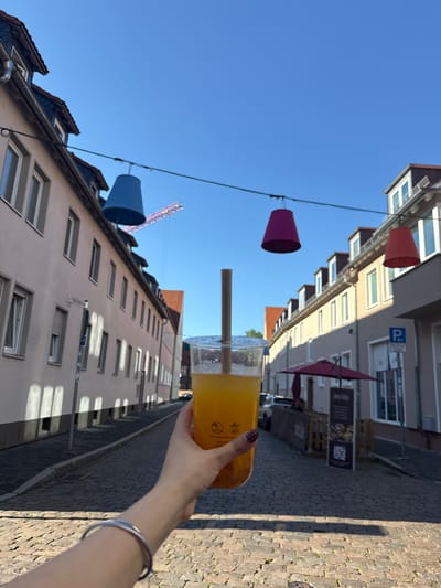 A street scene in Hanau, Germany, is captured from a low angle, with a focus on a hand holding a plastic cup filled with an orange beverage and a straw. The cup obscures some of the background. Overhead, colorful decorative cone-shaped lights hang suspended above the street. The buildings on either side of the street are multi-story structures with windows and varying rooflines. A construction crane is faintly visible in the background. A street sign is partially visible on the lower building, while a parking sign is visible on the higher building. The street is paved with cobblestones.