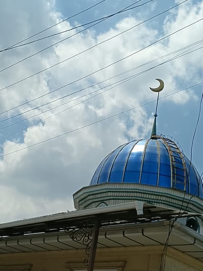 The image shows the blue dome of a mosque, topped with a crescent moon symbol, against a cloudy sky crisscrossed by power lines. A rusty ladder is attached to the side of the dome. The lower part of the image reveals the eaves of a building, with decorative metalwork and a portion of the building's facade visible. The scene is likely outdoors, possibly in the city of Osh, Kyrgyzstan, given the presence of a mosque. No people are visible in the frame.