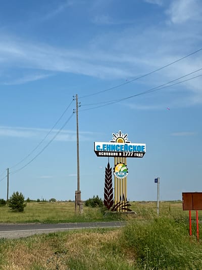 An outdoor, eye-level, medium shot shows a sign for the city of Eniseiskoe, Russia, on a bright, sunny day with a blue sky and wispy clouds. The sign, located in the foreground to the right, is tall and features the words "С.ЕНИСЕЙСКОЕ" (S. ENISEISKOE) in large, blue, capital letters with a stylized sun image above. Below the city name, in smaller text, it reads "ОСНОВАНО В 1777 ГОДУ" (FOUNDED IN 1777). The sign also includes a circular emblem with the letters "АПК" and smaller text that appears to be "ЕНИСЕЙСКОЕ." The sign is mounted on a structure resembling stalks of wheat.
To the left of the sign, a tall wooden utility pole with several wires stretches across the frame against the sky. Further to the left, another similar pole is visible in the distance. A paved road runs across the lower portion of the image, bordered by green grass and vegetation. The background consists of a large, flat, grassy field with scattered trees and some distant structures. Two small pink kites are visible flying in the sky, one to the left and one to the right of the main sign. To the far right of the sign, a brown rectangular sign with red posts is partially visible. The overall environment appears rural.