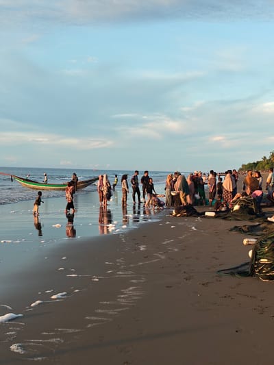 The image captures a bustling scene on the sandy shore of Uleekareung, Indonesia, at what appears to be sunset or early evening, given the soft lighting and pastel hues in the sky. A long, narrow fishing boat, painted green and red, rests partially in the shallow water, its reflection shimmering on the wet sand. Numerous people, a mix of adults and children, are gathered on the beach. Some are clustered together, seemingly engaged in conversation or activity, while others are more dispersed. Several individuals, particularly women, are wearing headscarves and traditional attire. Fishing nets, some piled in bundles and others spread out, are a prominent feature on the sand, indicating ongoing or recent fishing activities. Small waves gently lap at the shore, leaving trails of white foam. The overall atmosphere is one of community engagement with the sea, likely related to the fishing industry. The horizon shows a distant coastline with palm trees, suggesting a tropical or subtropical location.