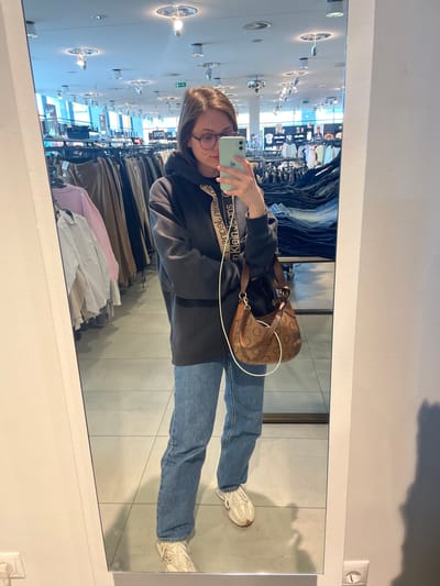 A young woman stands in front of a mirror in a clothing store, taking a selfie with her light green smartphone. She is wearing a dark grey hoodie with the words "Calvin Klein Jeans" in white on the straps, light blue jeans, and white sneakers. She is also carrying a brown handbag with a monogram pattern. The mirror reflects the store interior, including racks of clothing in various colors and styles, overhead lighting fixtures, and signage in the background. The store is located in Keszthely, Hungary.