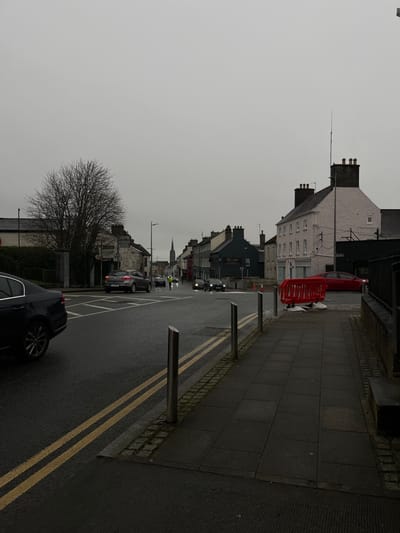 The scene depicts a street in Tullamore, Ireland, under an overcast, grey sky. The asphalt road surface appears wet. A dark paved sidewalk, edged by a stone curb and several silver-colored bollards, runs along the right side of the image. On this sidewalk, a red plastic temporary barrier is positioned, behind which a red sedan is parked alongside a building.
The roadway contains multiple vehicles in motion. A dark sedan is partially visible in the immediate foreground on the left. Further down the street, a grey SUV is observed in an intersection area. Several other dark vehicles with illuminated headlights are present in the middle distance. One individual wearing a high-visibility yellow vest stands near the center of the road, appearing to be engaged in traffic direction or roadside work.
A prominent, bare-branched deciduous tree is located on the left side of the street, obscuring part of a low building. A row of two and three-story buildings lines both sides of the street, exhibiting various architectural styles and colors, including a light pink building on the right and darker structures further down. A sign for "DELI'S" is visible on one building. In the far distance, a tall, slender church spire rises above the skyline. Double yellow lines mark the center of the visible road lanes.