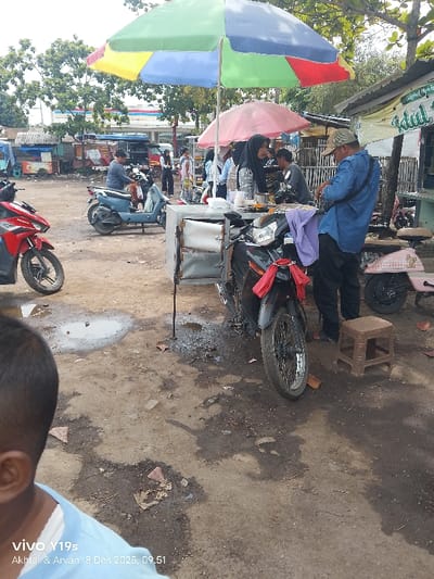 A marketplace scene shows a vendor in a blue shirt standing near a motorcycle with an attached food cart under a colorful umbrella. A customer wearing a headscarf stands nearby. A second umbrella shades the vendor. Other people and motorcycles fill the background, along with shops and other street vendors. A child's head and shoulder are visible in the foreground. The ground is uneven, showing dirt and small puddles.