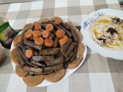 Two plates of Middle Eastern food rest on a beige and white checkered tablecloth. One plate is piled high with what looks like stuffed grape leaves, topped with sliced carrots and surrounded by peeled potatoes. The second plate contains a creamy, light-colored spread, possibly hummus, adorned with olive oil and dark spices. A bottle with a green label is partially visible in the background. The overall scene portrays a homemade, appetizing meal.