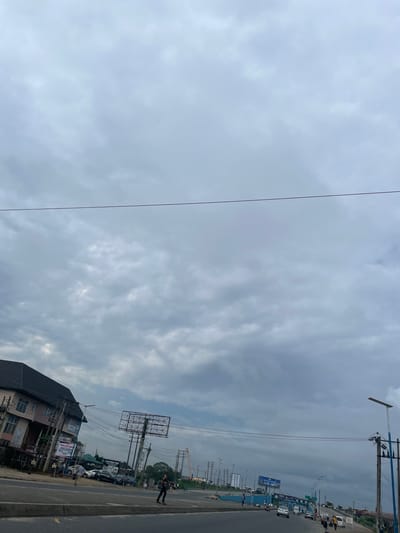 This image captures an overcast daytime scene of a busy roadside in Rumuosi, Nigeria. The photograph is taken from a slightly elevated perspective, looking across a multi-lane road towards buildings and infrastructure on the opposing side.

The sky is entirely covered with thick, grey, textured clouds, suggesting overcast weather and diffuse lighting. Numerous utility poles with a dense network of overhead wires dominate the visual space, running parallel to the road. A prominent feature on the left side of the road is a large, empty metal billboard structure. Below it and to the left, several multi-story buildings with dark roofs are visible, interspersed with smaller signboards. One white signboard clearly displays the text "OPPORTUNITY FOR YOUTH," and another sign further down reads "PLAYFIELD PARK."

The road itself shows moderate traffic, with various vehicles including cars, a white mini-bus or van, and what appears to be a construction vehicle or tractor further in the distance. Pedestrians are also visible along the roadside; one person in dark clothing stands near the right edge of the image, and others can be seen further down the road. The overall setting is consistent with a bustling urban or peri-urban environment, characterized by commercial activity, transportation, and dense infrastructure. No specific event is discernible, but it depicts a typical day in the area under cloudy skies.