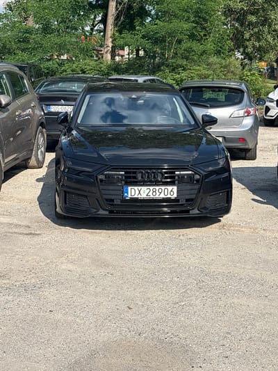 The image shows a black Audi A6 sedan parked on a gravel lot. The car's license plate is visible and reads "DX 28906". There are other cars parked in the background, including a silver Mazda and a grey SUV. Trees and some buildings are visible in the background. The location is identified as Kobyla Góra, Poland. The scene appears to be a sunny day.