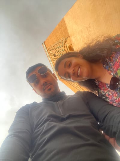 The image depicts a selfie of a man and a woman in Mechouar Kasbah, Morocco. The man is wearing sunglasses and a dark shirt. The woman is wearing a floral-patterned shirt and has sunglasses resting on her chest. Behind them is a building with brick and intricate tile work. The sky is overcast.