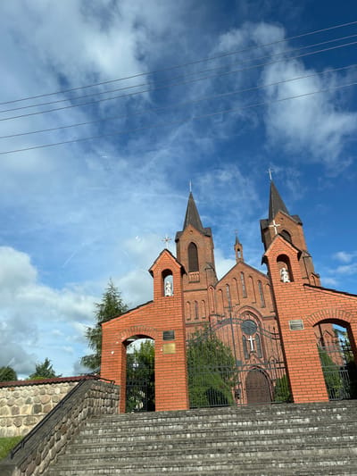 The image shows a red brick church with two spires against a blue sky with scattered clouds. In the foreground, there is a stone wall and a set of concrete stairs leading up to the church. The church has a large wooden door and several windows. There are also two statues of figures in niches on the facade. Electrical wires are visible in the upper part of the image. The church appears to be located in the city of Myory, Belarus.