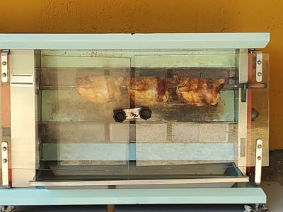 A rotisserie chicken cooker is positioned outdoors against a textured yellow wall. The cooker has a pale blue exterior and a glass front. Three chickens are rotating on a spit inside the cooker. The chickens are golden brown and appear to be cooked. The scene is captured from a frontal, eye-level perspective. The location is Cadalso, Spain.