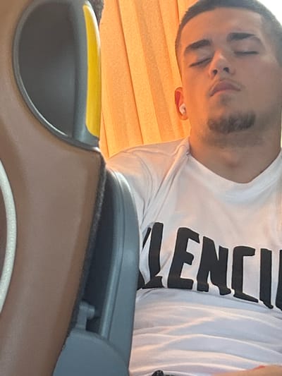 A young man with closed eyes and earbuds in his ears is seated next to a bus seat, possibly sleeping. He is wearing a white t-shirt with black lettering. The background consists of orange curtains and the interior of a bus. The image is cropped and close-up, focusing on the man and his immediate surroundings. The location is specified as Diadovo, Bulgaria.