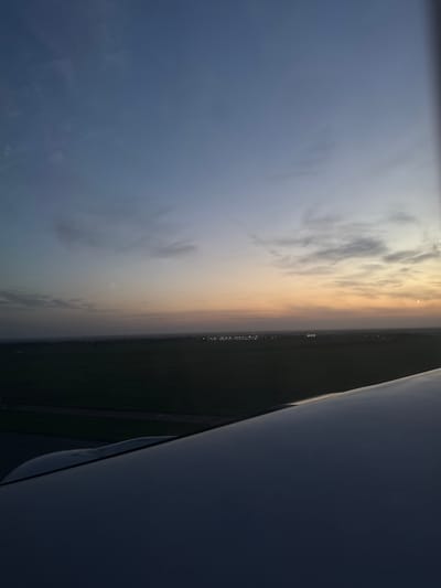 The image, taken from an aircraft in the vicinity of Stolbishche, Russia, presents a view of the horizon at twilight. The sky is a gradient, transitioning from a deep blue to a pale orange at the horizon line, marked by scattered clouds. Below the horizon is a dark expanse of land, punctuated by strings of lights suggestive of a road or urban development. The wing of the aircraft is visible in the foreground, partially obscuring the view.