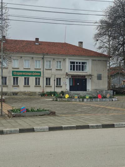 The image depicts a three-story building in Debnevo, Bulgaria. A green sign on the building reads "120 години читалище в Дебнево" (120 years community center in Debnevo). The building has a red-tiled roof and multiple windows. A sidewalk runs alongside the building, adorned with colorful, round decorations. A road is visible next to the sidewalk. Power lines run in front of the building and bare trees are to the left and in the background. The sky is overcast.