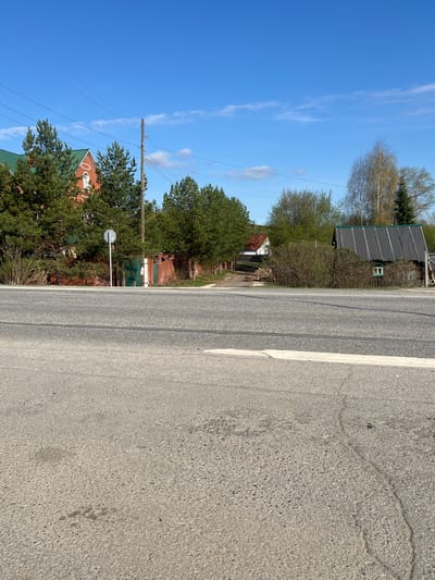 The image presents a road, likely paved, running parallel to a row of houses and trees. The sky is blue with some wispy clouds. A white line marks the edge of the road. There is an electricity pole visible with power lines. Based on the information given, the location is Yugo-Kamskiy, Russia. No people, actions, or interactions are visible.