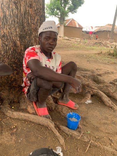 A young Black male, appearing to be in his late teens or early twenties, is crouched next to the base of a large tree, in what appears to be the city of Dull A, Nigeria. He is wearing a white beanie with the word "BALEN" printed on it in black lettering, a white t-shirt with red and pink abstract patterns, and dark shorts. He has on red slide sandals. His knees are up and his hands are resting on his knees. To his right, a small blue bucket contains a light-colored liquid or substance. The ground is dirt and is covered with tree roots. In the background, several rustic buildings with thatched or corrugated metal roofs are visible, suggesting a rural or semi-rural setting within Dull A, Nigeria. The sky is overcast.