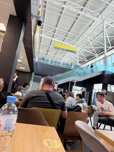 This image depicts a busy, modern indoor public space, likely an airport terminal or a large waiting area, in Тагашево, Russia. The perspective is slightly elevated, looking down and across a multi-level layout.
Numerous individuals are scattered throughout the scene, primarily seated in various chairs and at wooden tables. Most appear to be adults or young adults, dressed in casual attire. Activities largely involve waiting; many people are looking down, possibly at phones, while others are resting or observing their surroundings. A man in a grey cap is seen from behind in the foreground, and further back, a person wearing an Adidas-branded white shirt sits at a wooden table.
The setting is characterized by its contemporary architecture, featuring high ceilings supported by an intricate network of white structural trusses and corrugated metal panels. Large glass barriers and dark, sleek facades delineate different levels and sections of the building. Ample natural light illuminates the space, suggesting it is daytime. A security camera is mounted on one of the dark structural elements.
Visible text provides location and directional information. A prominent yellow sign, partially seen at different points in the image, displays directional arrows along with "DUTY FREE" in English, "2 ЭТАЖ" (meaning "2nd floor" in Russian), and "ТАГАШЕВО" (the specified location). Other sections of the sign show numbers "5.6" and "7" with arrows, likely indicating gate numbers or other directions. On a table in the foreground, a yellow circular sticker bears the Cyrillic text "УВАЖАЕМЫЕ ПАССАЖИРЫ!" (meaning "Dear Passengers!"). A bottle of Evian water is also visible on a table in the foreground.
The overall atmosphere is one of calm transit, typical of a busy travel hub.