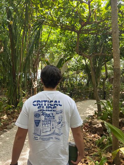 The image shows a young man wearing a white t-shirt with blue lettering, standing on a white gravel path in a lush, tropical forest. The t-shirt depicts a cartoon illustration of a vending machine called "Wave $5 Machine" with a surfer standing beside it, and the words "Critical Slide" at the top. The man is holding a stainless steel water bottle in his right hand. The forest is thick with tall trees and dense foliage. The path leads to a clearing. The coordinates (latitude: 9.635, longitude: -85.161) place this location in a residential area just east of Santa Teresa, Costa Rica.
