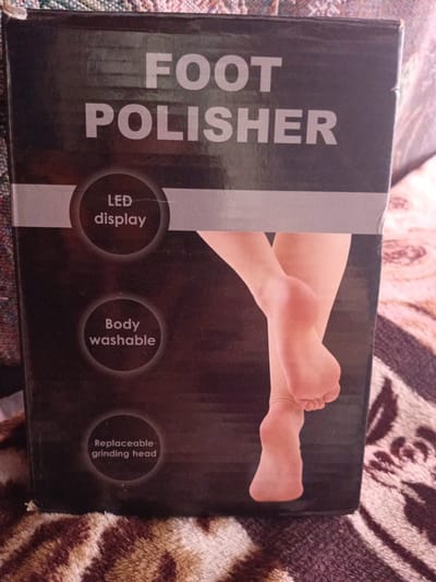 The image shows the front of a black box for a foot polisher. The text on the box reads "FOOT POLISHER" in large, white, capitalized letters. Below this, there are three circular icons with white text inside. The top icon reads "LED display." The middle icon reads "Body washable." The bottom icon reads "Replaceable grinding head." A portion of a bare foot and lower leg is visible on the right side of the box. The box is lying on a patterned surface, possibly a blanket or rug, which has shades of brown and white. The background is out of focus and appears to be a wall. There is no indication that this image was taken in Monastirische, Russia.
