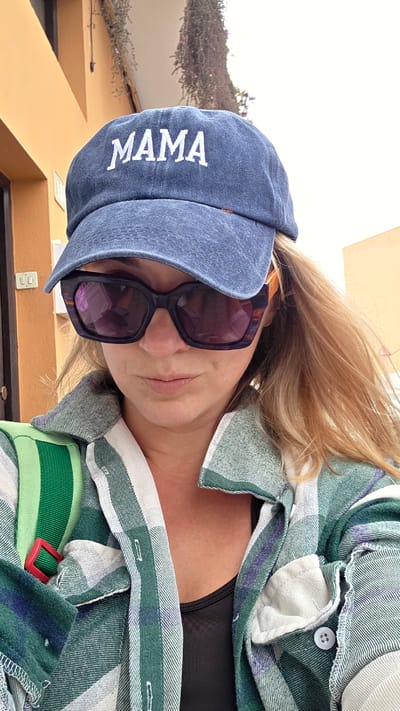 A medium shot shows a woman wearing a blue baseball cap with "MAMA" embroidered in white, large dark-rimmed sunglasses, and an open green, white, and purple plaid shirt over a black top. A green and white striped strap with a red buckle crosses her chest, suggesting a bag or backpack. Her blond hair is visible on the right side of her face. The background consists of an orange building to the left and a light-colored wall to the right, with some greenery hanging from above. The scene appears to be outdoors, possibly on a street or walkway in Guía de Isora, Spain, indicated by the architectural style.