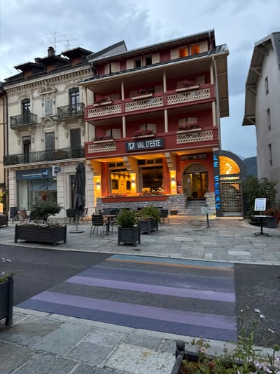 The image depicts a street scene in Saint-Gervais-les-Bains, France, featuring the Hotel Val d'Este and Le Serac restaurant. The hotel is a prominent red building with multiple balconies, some adorned with flower boxes. A sign for "VAL D'ESTE" is visible on the hotel facade. Adjacent to the hotel is a bank, "BANQUE POPULAIRE +BX". In front of the businesses, there are outdoor seating areas with tables and chairs. A large grey umbrella is propped up near the bank. The foreground shows a crosswalk painted with purple, blue, and orange stripes. The sky is overcast, suggesting it is either early morning or late evening. The architecture is typical of a European mountain town, with older, ornate buildings alongside modern establishments.