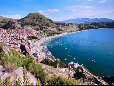 The image shows a panoramic view of a coastal city nestled at the base of rolling green hills, with a vast body of turquoise water stretching to the horizon. The city, characterized by its densely packed buildings with red-tiled roofs, extends along the shoreline. Numerous boats are visible in the water, both docked along the coast and scattered across the open expanse. The foreground is dominated by rocky terrain with sparse vegetation, suggesting a natural, somewhat rugged landscape overlooking the developed area. In the distance, a range of hazy blue mountains forms the backdrop against a bright sky with scattered clouds. The scene appears to be captured from an elevated viewpoint, providing a sweeping perspective of the city, its harbor, and the surrounding natural environment.
