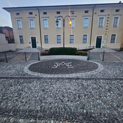 The image shows a building with the word "ORATORIO" written vertically on its facade in Calcinate, Italy. The building is light yellow and has several windows with white frames and dark grates. In front of the building, there is a cobbled courtyard. A lamppost with two lights, one lit orange, stands in the middle of the courtyard. A circular mosaic made of white and dark grey pebbles is visible on the ground. There are also stairs leading up to a green door of the building. The sky is overcast.