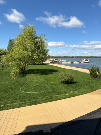 A bright, wide-angle photograph captures a serene lakeside scene on a sunny day in Bil'dino, Russia. The foreground is dominated by a light-colored wooden deck, its planks laid in a curved pattern, with a large expanse of meticulously manicured green lawn stretching behind it. A mature, leafy willow tree stands prominently on the lawn, its branches casting a shadow. Further back, a body of water, likely a lake, reflects the blue sky dotted with fluffy white clouds. Along the water's edge, a yellow wooden boardwalk leads to a series of floating docks, where a small white boat is moored. The distant horizon is lined with trees, creating a picturesque natural backdrop. The scene conveys a sense of tranquility and recreational potential.