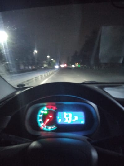 The image depicts the interior of a car at night, presumably in Urtaaul, Uzbekistan based on the prompt. The dashboard and steering wheel are visible in the foreground. The speedometer displays a reading, likely indicating the car is in motion. Through the windshield, a road is visible with streetlights lining the side. Other vehicles are suggested by the presence of headlights. The environment appears urban or suburban, judging by the presence of streetlights and illuminated buildings. There are no people visible in the photograph.
