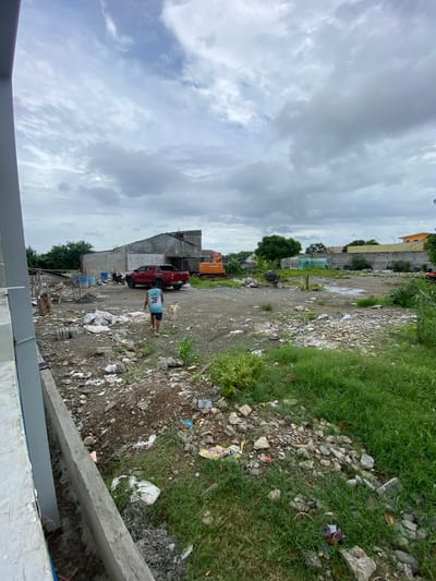 This is a vertically oriented, outdoor photograph of a somewhat neglected urban landscape, likely in San Jose del Monte, Philippines. The scene appears to be an undeveloped or under-construction lot, characterized by dirt paths, scattered debris, rocks, and patches of overgrown grass. In the mid-ground, a person wearing a blue and white jersey with the number 28 is walking. Beside them, a small, light-colored dog is also present.

Several vehicles are visible: a red pickup truck is parked near a low-rise building. Further in the background, an orange construction vehicle is present. The buildings in the background are varied, with some appearing to be unfinished or in disrepair, featuring concrete walls and corrugated metal roofing. One building has graffiti on its side. There is also a sign that appears to say "LOT FOR LEA" with other unreadable text below it.

The sky is overcast with heavy, grey clouds, suggesting it is a cloudy day, possibly leading up to or during rain. The overall impression is one of a transitional space, possibly an area undergoing development or facing neglect, with signs of human activity interspersed with the natural elements and refuse. The lighting is diffused due to the cloudy conditions.