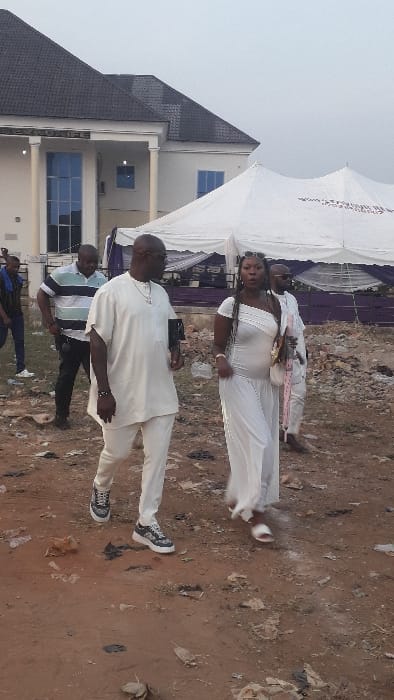An outdoor scene in Agukwu Nri, Nigeria, depicts six individuals navigating an unpaved, reddish-brown dirt area. The ground is uneven and littered with small rocks, dry leaves, and discarded debris. In the background, to the left, stands a light-colored, two-story building with a dark gabled roof, prominent white columns, and blue-tinted windows. A partially visible sign above the entrance reads "OBEKUNIFE". To the right, a large white event canopy with purple interior draping is visible, bearing partial text "WWW.ATAYOUTU.COM" and "2023". The sky is overcast.
In the foreground, a dark-skinned, bald male wearing dark-rimmed glasses, a short beard, a light-colored traditional outfit, gold chain, dark bracelet, and black-and-white sneakers walks forward, holding a dark object in his left hand. Beside him, a dark-skinned female with long braided hair, wearing sunglasses on her head, a one-shouldered light-colored long dress, light-colored sandals, and a light-colored purse, also walks forward with her mouth slightly open.
Behind these two figures, a third dark-skinned male in a light blue and white striped polo shirt and dark trousers walks to the left. Further behind him, a fourth dark-skinned male in a light-colored traditional outfit and sunglasses walks to the right. To the far left in the background, a fifth dark-skinned male in a dark t-shirt and blue jeans stands partially visible. A sixth male is partially visible behind the polo-shirted individual. The primary action involves the group walking from left to right across the frame.