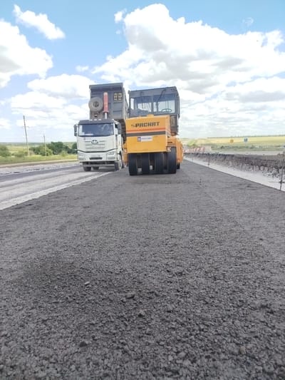 In Komyshuvate, Ukraine, a road construction scene is depicted. The foreground is dominated by a freshly laid asphalt surface, textured and dark gray. In the middle ground, a large yellow road roller with multiple drums is actively compacting the asphalt. To the left of the roller, a white dump truck is positioned, its cargo bed raised, suggesting it has recently deposited material. The truck's license plate, though partially obscured, appears to have Cyrillic characters. Behind the construction equipment, the landscape transitions to a rural setting with fields and scattered trees under a bright, cloudy sky. The scene captures a moment of infrastructure development.