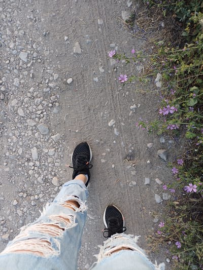 The image shows a person's legs from the knees down, wearing ripped light-wash denim jeans and black athletic shoes with white socks. The person is standing on a dirt path with scattered gravel and small stones. To the right of the path, there is a patch of green vegetation with small purple flowers and dry grass. The scene is outdoors, likely in a natural setting or a park. There is no specific indication of the city of Medovina, Bulgaria within the visual information of the image.