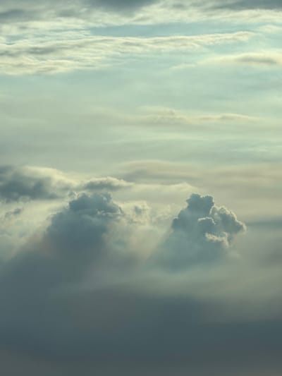 The image depicts a dramatic cloudscape from an aerial perspective, possibly taken from an airplane. The sky is filled with layers of clouds, ranging from soft, wispy formations at higher altitudes to denser, darker masses closer to the viewer. Sunlight filters through the clouds, creating a sense of depth and texture. Shadows are cast by the clouds, adding to the visual drama. The overall color palette is muted, with shades of gray, blue, and pale yellow dominating the scene. There are no discernible human elements or man-made structures visible. The image does not provide any specific information or context to place it in the city of Tort-Kuduk, Kazakhstan, or any other location.