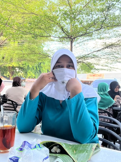 A medium shot captures a young Indonesian woman wearing a white hijab and a teal top, seated outdoors at a table with a glass of iced tea and food wrapped in banana leaves. She is adjusting her white face mask. In the background, other individuals are visible, also wearing hijabs, and there appears to be a street scene with a car and signage. The setting suggests a casual dining environment in Keuniree, Indonesia, under the shade of leafy trees.