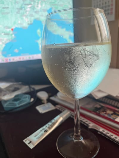 In the foreground, a clear wine glass filled with a pale yellow liquid and ice cubes is positioned on a dark, reflective surface. Condensation beads cover the exterior of the glass, indicating it is cold. Behind the glass, a computer monitor displays a map with a red marker, possibly indicating a location within Torre Pacheco, Spain. Several small objects, including a tube of what appears to be lip balm or medication, a small white container, and what looks like a sanitizing wipe packet, are scattered on the surface. In the background, a calendar is partially visible. The overall environment suggests a desk or workspace.