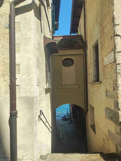 The image depicts a narrow alleyway in Orta San Giulio, Italy, leading towards a body of water. The alley is flanked by aged, textured buildings made of stucco and stone. On the right side of the alley, a white sign with the text "VICOLO S.DEMETRIO" is affixed to the building. A dark, vertical downspout runs along the left building. The alley opens up to a view of a bright blue body of water with a small, dark boat docked at its edge. The ground of the alley is paved with cobblestones. The scene is illuminated by bright sunlight, casting strong shadows. There are no people visible in the image.