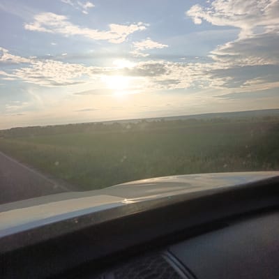 The image shows a landscape scene from the perspective of a vehicle's dashboard. In the foreground, the upper portion of a car's dashboard is visible, with a smooth, light-colored surface reflecting the sky. Beyond the dashboard, a road runs along the left side of the image, bordered by a field of tall, green grass. In the middle ground, a treeline stretches across the horizon, with a large field of what appears to be cultivated land behind it. The background is dominated by a vast sky filled with scattered white clouds. The sun is visible in the upper center of the sky, casting a bright light and creating a hazy effect over the landscape. The overall scene suggests an outdoor setting, possibly in a rural area, during daylight hours.