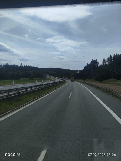 The image shows a highway scene on a cloudy day. The highway is asphalt with white lane markings. There are guardrails and white posts along the side of the road. A dense forest lines the right side of the highway, and a field of crops is visible on the left. Several trucks are present on the highway, some in motion and some appearing to be stopped in traffic. The timestamp in the bottom right corner indicates the image was taken on July 7, 2026, at 15:06. The bottom left corner displays "POCO F5". The scene is consistent with a section of the A93 highway near Köditz, Germany, given the presence of a multi-lane highway bordered by forest and agricultural land. There are no people clearly visible in the image.